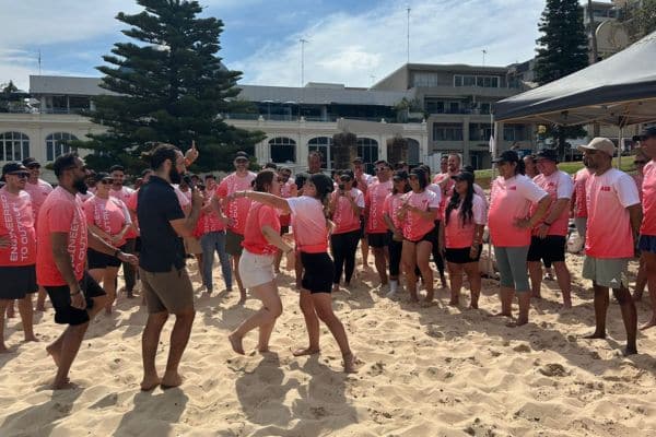 ABB corporate team in matching pink t-shirts receiving instructions on the beach during a Mini Beach Olympics team building event with Pinnacle Team Events