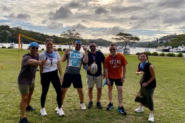 Corporate team posing with medals and a rugby ball on a waterfront lawn after completing a Mini Olympics outdoor team building event with Pinnacle Team Events