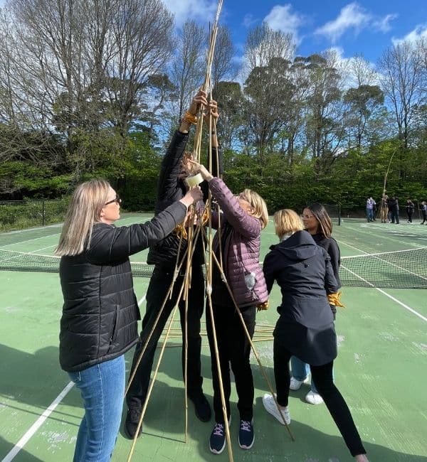 Four participants collaborating to build a tall bamboo pole structure on a tennis court during The Scramble team building activity