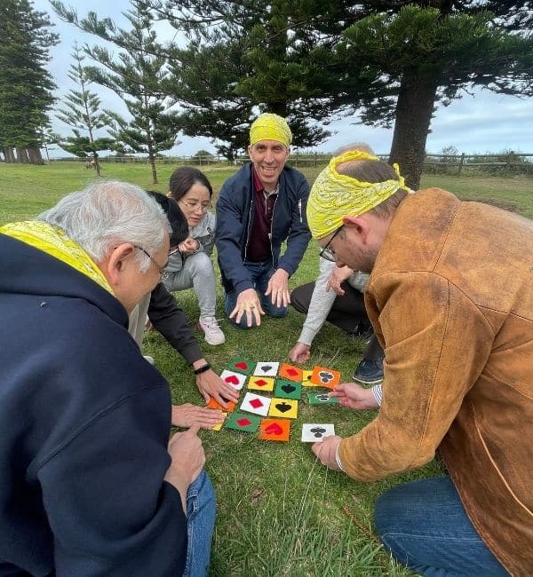 Four participants wearing yellow bandanas kneeling on grass to solve a playing card suit tile puzzle during The Scramble team building activity