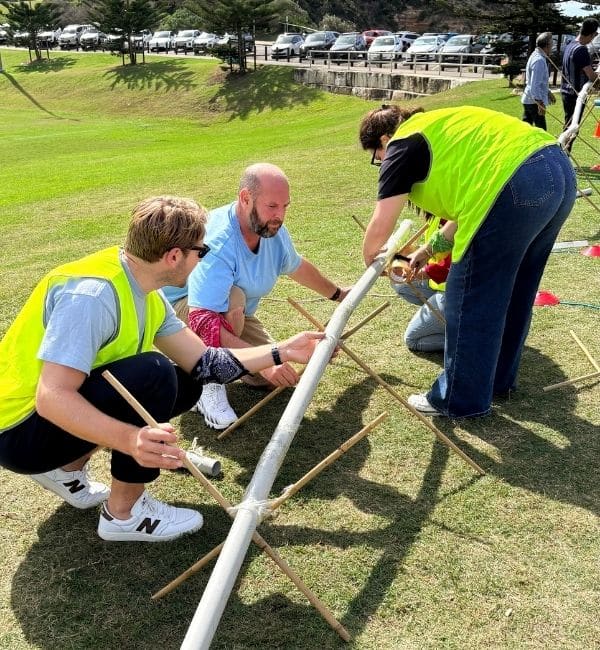 Three participants in hi-vis vests crouching on grass to connect PVC pipe sections supported by bamboo tripods during the Project Pipeline team building activity