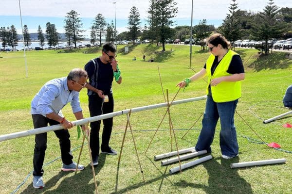 Three participants assembling a PVC pipe structure with bamboo supports on a coastal grass reserve during the Project Pipeline team building activity