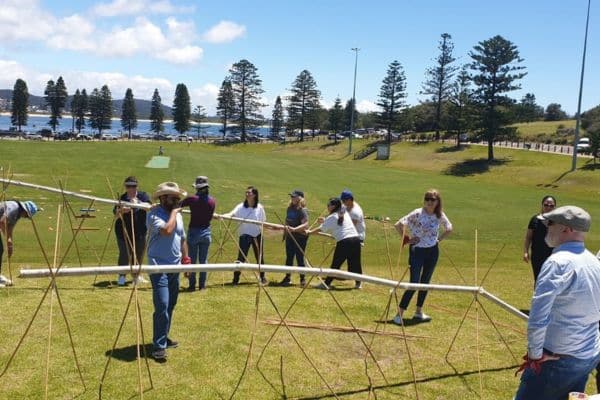 Three participants assembling a PVC pipe structure with bamboo supports on a coastal grass reserve during the Project Pipeline team building activity