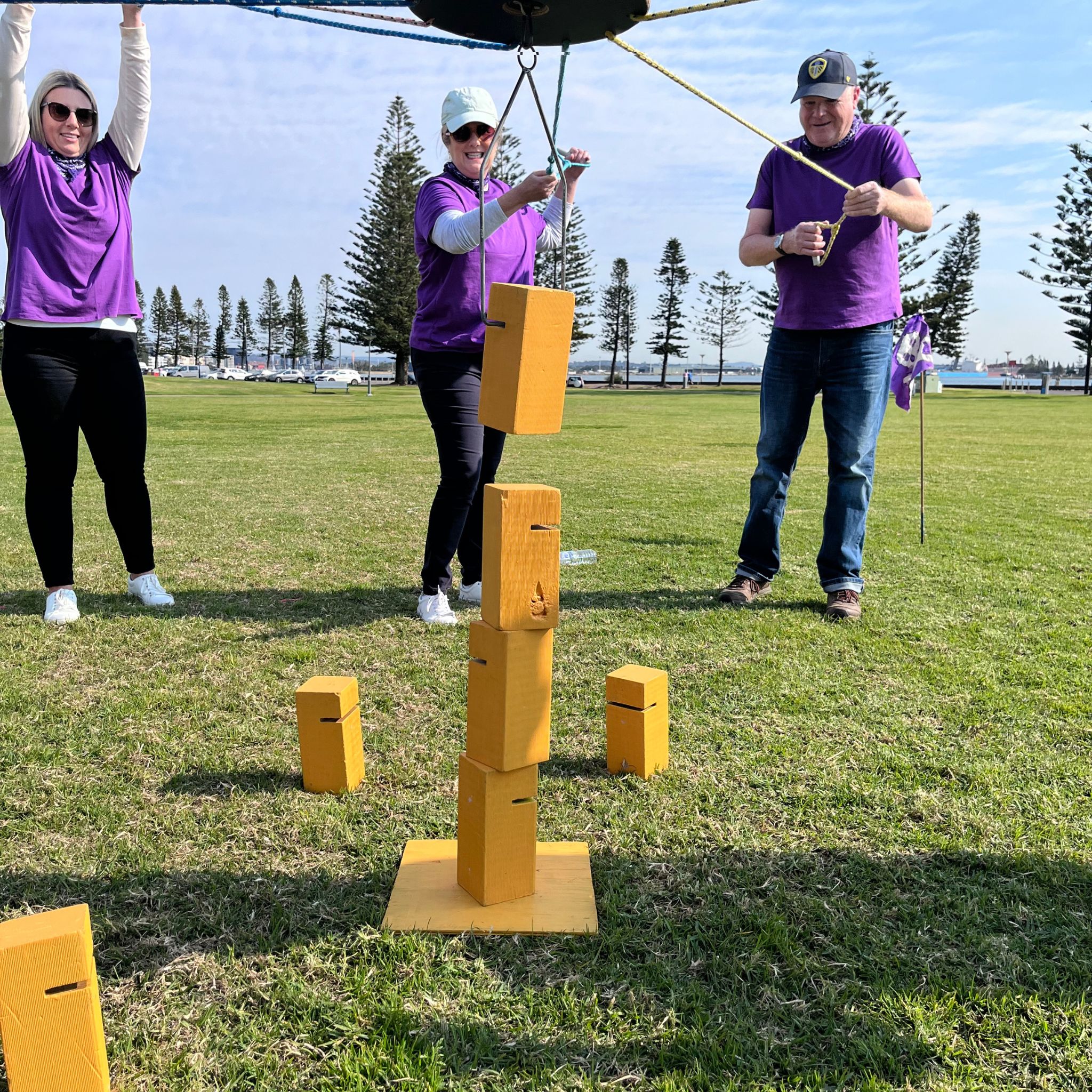 Corporate team in purple t-shirts successfully stacking blocks using a rope and pulley system during the Skyscraper challenge at a Ransom outdoor team building event with Pinnacle Team Events