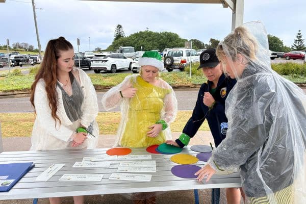 Four participants in rain ponchos working through a Scramble challenge at an outdoor table with coloured discs and clue cards