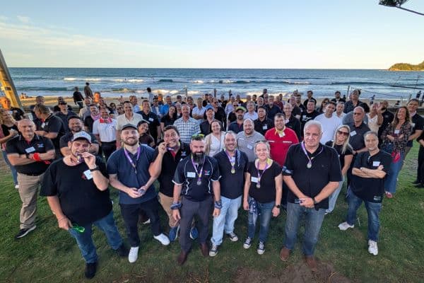 Large group of Scramble participants posing for a group photo on a grassy beachfront at golden hour