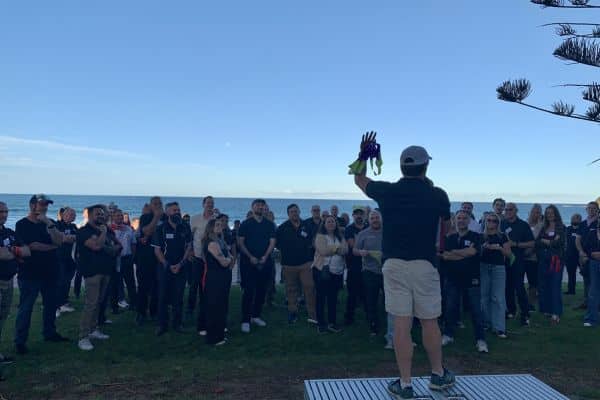 Facilitator addressing a large group of participants on a grassy beachfront during The Scramble team building event