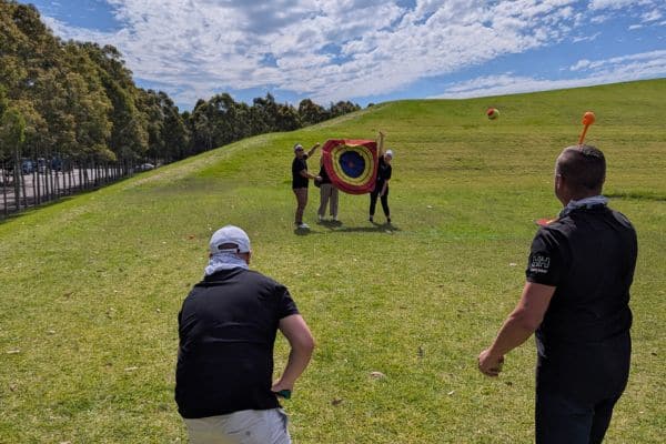 Corporate team members throwing a ball at a target held by teammates on a grass hill during a Treasure Hunt outdoor activity with Pinnacle Team Events